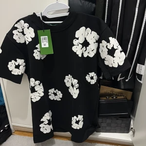 Svart blommig t-shirt från Denim Tears - Svart t-shirt från Denim Tears med stora vita blommor tryckta över hela plagget. Oversized fit och klassisk rund hals. Perfekt statement piece för dig som vill sticka ut med en unik och trendig look.