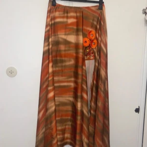 Lång kjol i siden med orange detaljer - Unik långkjol i mjukt siden med vågiga ränder i orange, brunt och beige. Kjolen har en handgjord applikation med orange blommor och glittriga stenar framtill. Flödande passform och lätt glansigt tyg som ger en bohemisk vibe.