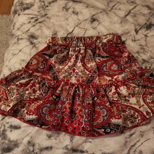 Röd mönstrad kortkjol från SHEIN - Supersöt kortkjol från SHEIN i storlek XS med färgstarkt paisleymönster i rött, beige, svart och blått. Kjolen har volangkant och resår i midjan för en bekväm passform. Perfekt för dig som vill sticka ut med en bohemisk vibe.