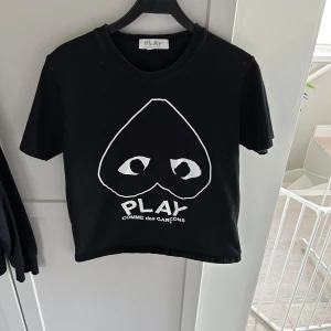 Svart t-shirt från Comme des Garçons Play - Svart t-shirt med stort vitt hjärtmotiv och ögon på bröstet samt texten 'PLAY COMME des GARÇONS' under. Klassisk rund hals och korta ärmar. Perfekt statement-plagg för dig som gillar streetwear.