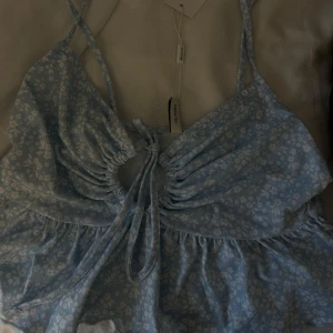 Ljusblå blommig linnetopp från NLY Trend - Supersöt ljusblå linnetopp med små vita blommor från NLY Trend. Toppen har smala axelband, knytning framtill och en lätt volang nertill. Perfekt för varma dagar och ger en romantisk känsla.