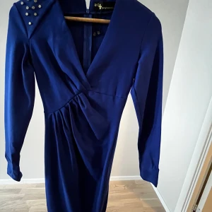 Kornblå klänning med nitar från Arzu Kaprol - Säljer en kornblå långärmad klänning från Arzu Kaprol med v-ringning och dekorativa nitar på ena axeln. Klänningen har omlott-detalj framtill och markerad midja för en snygg siluett.