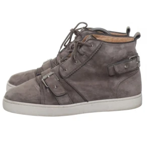  Christian Louboutin skor - Säljer ett par grå höga sneakers från Christian Louboutin i mocka med snörning och spänndetaljer. Skorna har rund tå, vit platt sula och diskret logga på sidan. Perfekta för dig som vill ha något unikt och stilrent.