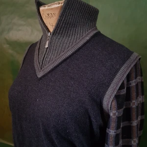 Mörkgrå stickad tröja med dragkedja - Säljer en mörkgrå stickad tröja med hög ribbad krage och dragkedja framtill. Ärmarna har rutigt mönster i grått och blått, vilket ger en unik look. Perfekt för dig som gillar stilrena detaljer och vill ha något annorlunda.