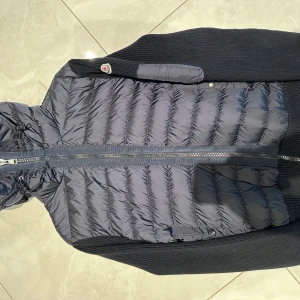 Mörkblå pufferjacka från Moncler - Snygg mörkblå pufferjacka/ cardigan från Moncler med stickade ärmar och huva. Jackan har dragkedja framtill, fickor med knappar och en extra ficka med dragkedja på ärmen. Perfekt för kyliga dagar och ger en sportig look pris 5500 kom med bud då kvitto o allt medkommer.
