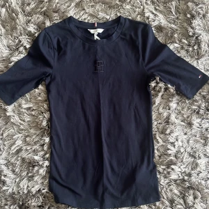 Mörkblå topp från Tommy Hilfiger - Säljer en mörkblå topp från Tommy Hilfiger med broderad TH-logga på bröstet. Toppen har korta ärmar och rund halsringning. Perfekt för en stilren och klassisk look.