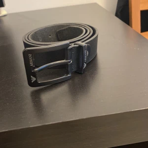 Svart läderbälte från Armani - Snyggt svart bälte från Armani i äkta läder med klassiskt spänne och diskret logga på metallspännet. Perfekt accessoar för att lyfta din outfit med en stilren touch.