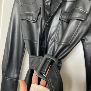 Svart skinnjacka med bälte från Zara - Säljer en svart skinnjacka från Zara med bälte i midjan och klassisk krage. Jackan har långa ärmar, två bröstfickor med lock och knappar framtill. Perfekt för dig som vill ha en stilren och edgy look.