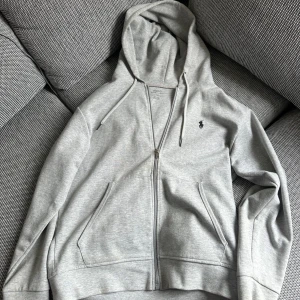 Polo Ralph Lauren zip-hoodie grå - Stilren och klassisk grå zip-hoodie från Polo Ralph Lauren. Broderad logga på bröstet, fickor framtill och justerbar huva med snören. Tröjan är i fint skick utan några defekter. Skön och mjuk bomullskvalitet – perfekt för vardagsbruk eller lager på lager. Passar både dam och herr.