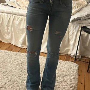 Lågmidjade blå bootcut jeans med slitningar från Lee - Säljer ett par blå bootcut jeans från Lee med slitningar på låren och knäna. Jeansen har klassisk femficksdesign. Midjemått:36 cm Innerbenslängden:80 cm (har två små fläckar, skriv för bild) Hör av dig om du vill ha fler bilder eller har någon fråga❤️