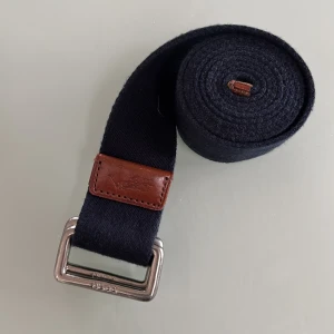 Mörkblått tygbälte från Polo - Snyggt mörkblått bälte i tyg från Polo med silverfärgat metallspänne och bruna skinn-detaljer. Perfekt accessoar för att lyfta din outfit med en klassisk touch. Bältet har en enkel och stilren design.