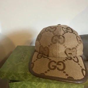 Beige keps från Gucci med GG-mönster - Snygg beige keps från Gucci med klassiskt GG-mönster i brunt. Kepsen har böjd skärm och är tillverkad i bomullsmaterial med detaljer i brunt runt skärmen. Perfekt accessoar för en trendig look.