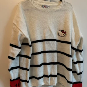 Vit och röd Hello Kitty stickad tröja - Supergullig stickad tröja från Hello Kitty i vitt med svarta ränder och röd detalj på ärmsluten. Broderad Hello Kitty-logga på bröstet. Perfekt för dig som älskar söta och unika plagg!