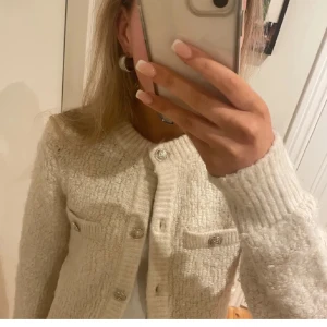 Vit stickad kofta med knappar - Mysig och fluffig vit stickad kofta med rund hals och dekorativa silvriga knappar framtill. Koftan har långa ärmar och två fickor på bröstet. Perfekt för lager-på-lager och enkel att styla till olika outfits.