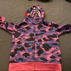 Lila och rosa camouflage hoodie med hajmotiv - Säljer en hoodie med dragkedja i lila, rosa och svart camouflage. Huvan har ett hajmotiv med tänder och ögon, och nederkanten är rosa. Perfekt för dig som vill sticka ut med en unik streetwear-stil.