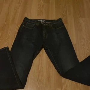 Mörkblå jeansbyxor från Grant - Säljer ett par klassiska mörkblå jeans från Grant med raka ben och kontrastsömmar. Byxorna har fem fickor och läderpatch bak i midjan. Perfekta för dig som gillar en tidlös stil.