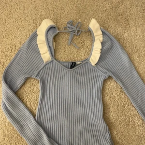 Ljusblå ribbad topp med volangdetalj - Supersöt ljusblå ribbad topp med långa ärmar och vit volang runt halsringningen. Knytning i nacken för extra detalj. Perfekt för dig som vill ha något unikt och trendigt i garderoben! 