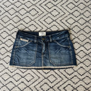 Lågmidjad jeanskjol med spetsdetalj  - Mörkblå jeanskjol med snygga fickor fram och bak och spetsdetalj längs kanterna. Klassisk femficksmodell med knapp och dragkedja framtill. Perfekt för dig som gillar denim med en söt twist. Spetsen är påsydd noggrant! 36 i midja rakt över 