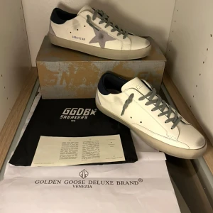 Golden Goose skor storlek 43 - Golden Goose Superstar sneakers i vitt skinn med grå stjärndetalj på sidan, grå snörning och mörkblå häl. Klassisk låg modell med platt sula och rund tå. Kommer med original box och dustbag. Skornas skick är 10/10 