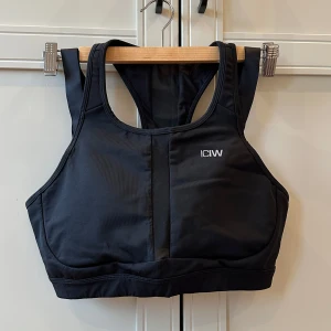 Svart sport-bh från ICANIWILL - Snygg svart sport-bh från ICANIWILL med brottarrygg och breda axelband. Perfekt för träning med bra stöd och stilren design. Liten logga framtill. Tillverkad i stretchigt syntetmaterial.