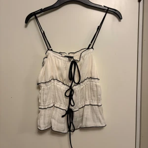 Vit och svart linnetopp från Gina Tricot - Säljer en söt vit linnetopp från Gina Tricot med svarta kontrastdetaljer och smala axelband. Toppen har volangiga lager och knyt framtill, vilket ger en lekfull och trendig look. Perfekt för varma dagar! Dessutom aldrig använd, prislapp sitter kvar.