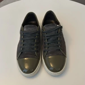 Grå sneakers från Lanvin med gröna detaljer - Säljer ett par grå sneakers från Lanvin med gröna lackdetaljer på tån och matchande gröna skosnören. Ovandelen är i mocka och sulan är vit. Klassisk rund tå och platt sula. Perfekta för dig som gillar stilrena och exklusiva sneakers.