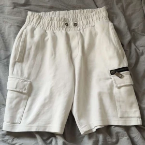 Vita cargo shorts från Moncler - Säljer ett par vita shorts från Moncler med elastisk midja och snörning. Shortsen har flera fickor, inklusive cargofickor på sidorna och en bakficka med Moncler-logga. Detalj med dragkedja på ena fickan. Perfekta för sommaren.