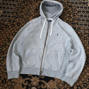 Hoodie från Polo Ralph Lauren - Klassisk grå hoodie från Polo Ralph Lauren med dragkedja och huva. Jackan har vita snören, två fickor framtill och en liten broderad logga på bröstet. Perfekt för en avslappnad stil.