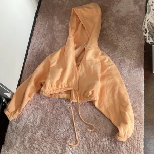 Persikofärgad croppad hoodie från H&M - Säljer en persikofärgad croppad hoodie från H&M med huva och dragsko i midjan. Jackan har dragkedja framtill och långa ärmar med resår vid ärmslut. Perfekt för en avslappnad och trendig look.
