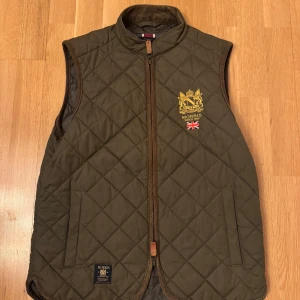 Olivgrön quiltad väst från Morris - Säljer en olivgrön quiltad väst från Morris med guldbroderad logga och brittisk flagga på bröstet. Västen har dragkedja framtill och två fickor. Perfekt för lager-på-lager och snygg till många outfits. Storlek XS, men jag är S och den passa mig. 