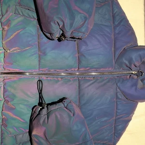 Iridescent pufferjacka med dragkedja - Cool pufferjacka i iriserande blå-lila nyans med dragkedja framtill och två stora fickor. Jackan har hög krage och quiltad design som ger extra volym och värme. Perfekt för dig som vill sticka ut i vinterkylan.