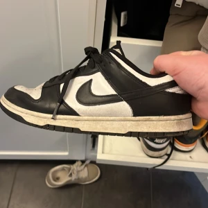 Nike Dunk Low svart/vit - Nike Dunk Low i svart och vitt med klassisk design. Skon har svart Swoosh, svarta snören och svart-vit ovandel i skinn. Yttersulan är svart och vit, och tån är rundad med perforeringar för ventilation. Perfekt för dig som gillar streetwear. Som du ser på bilderna så är de använda mycket