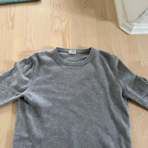 Grå stickad tröja från Asket - Säljer en klassisk grå stickad tröja från Asket. Tröjan har rund halsringning och långa ärmar med ribbade muddar. Perfekt basplagg i garderoben och passar till allt. Skitsnygg med en skjorta under eller bara en vanlig t shirt! Har använts mycket och har några slitningar men inget som påverkar hur den sitter eller hur snygg den är!