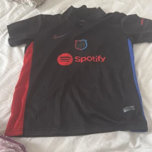 FC Barcelona svart fotbollströja med Spotify-logga - Säljer en svart FC Barcelona fotbollströja med röda och blå detaljer på sidorna. Tröjan har korta ärmar, klubbmärke och Spotify-logga på bröstet samt UNHCR-tryck på ryggen. Perfekt för dig som älskar Barca!
