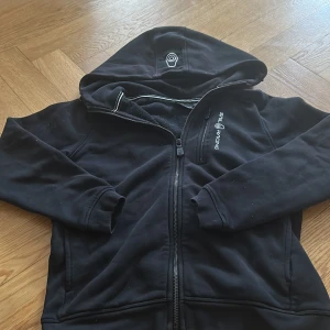 Svart ziphoodie från Sail Racing - Svart ziphoodie från Sail Racing med dragkedja framtill och broderad logga på bröstet. Jackan har huva och en ficka med dragkedja. Perfekt för en sportig och avslappnad stil.