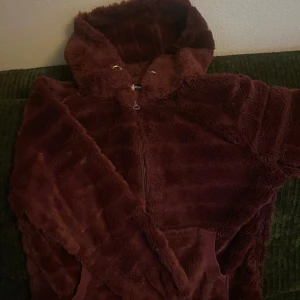 Vinröd teddyjacka med huva från Hunkemöller - Mysig vinröd teddyjacka från Hunkemöller med huva och dragkedja framtill. Jackan är fluffig och har en avslappnad passform, perfekt för kyliga dagar. Snygg och enkel att matcha med olika outfits.