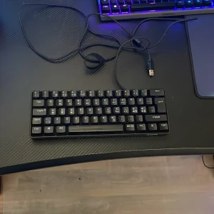 NOS C250 RGB Mekaniskt Tangentbord - Säljer ett kompakt NOS C250 RGB mekaniskt tangentbord med röda switchar och snygg RGB-belysning. Tangentbordet är svart, har en minimalistisk design och ansluts via USB. Perfekt för gaming eller skrivbordsarbete.