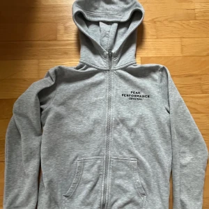 Grå hoodie från Peak Performance - Säljer en ljusgrå hoodie från Peak Performance med dragkedja och huva. Tröjan har broderad logga på bröstet och två fickor framtill. Perfekt för en avslappnad stil och enkel att matcha med det mesta.