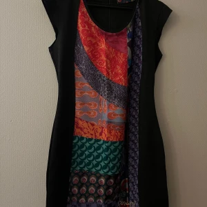 Färgglad mönstrad klänning med svarta sidor - Unik klänning med svart bas och färgglada mönster i rött, orange, grönt och lila framtill. Rund halsringning och ärmlös design. Klänningen har en figurnära passform och olika grafiska mönster som ger en cool och modern look.