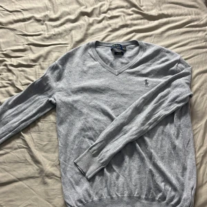 Grå v-ringad tröja från Polo Ralph Lauren - Säljer en stilren grå v-ringad tröja från Polo Ralph Lauren i mjuk pima bomull. Tröjan har lång ärm, ribbade muddar och den klassiska broderade loggan på bröstet. Perfekt för dig som gillar en slim fit passform. Som ny förutom att polo lappen är lös men går enkelt att sy tillbaka.
