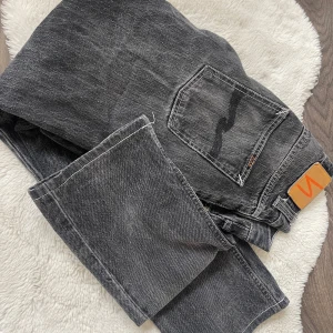 Grå jeansbyxor från Nudie Jeans - Säljer ett par grå jeansbyxor från Nudie Jeans med klassisk femficksdesign och raka ben. Byxorna har en snygg tvättad look och läderpatch bak i midjan. Perfekta för en avslappnad stil.