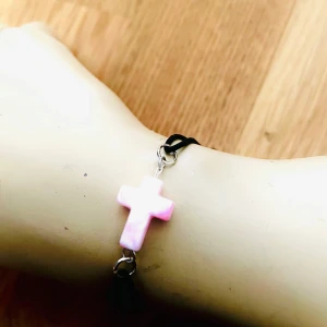By prisförslag- Armband med rosa kors - Säljer ett armband med svart band och ett ljusrosa kors i mitten. Krysset är gjort av ett glansigt material och sitter fast med små metallringar. Perfekt för dig som gillar enkel men snygg design.