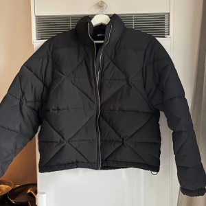 Svart pufferjacka med dragkedja - Säljer en svart pufferjacka med hög krage och dragkedja framtill. Jackan har ett quiltat mönster och är något croppad i modellen. Använd fåtal gånger, som ny.