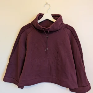 Puma - Cropped sweatshirt S/36 - Unik lila croppad hoodie från Puma med broderad logga framtill och mysiga teddy-detaljer längs ärmarna. Tröjan har hög krage med dragsko och är tillverkad i bomulll.