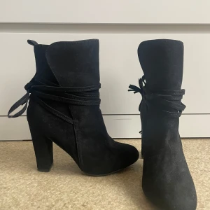 Svarta mocka boots med klack och knyt - Säljer ett par svarta boots i mocka med hög klack och snygga knytband runt ankeln. Skorna har rund tå och dekorativa tofsar baktill. Perfekta för att lyfta din outfit!