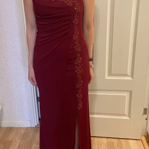 Vinröd långklänning med spetsdetaljer - Säljer en elegant vinröd långklänning med smala axelband och vacker spets längs ena sidan framtill. Klänningen har en slits framtill och figurnära passform, perfekt för fest eller bal. Materialet är mjukt och följsamt.passar 34-38, stretch kanske även 40.