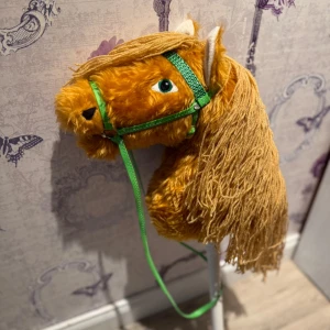 Käpphäst med långt garnman - Säljer en käpphäst med mjuk, ljusbrun päls och långt, gult garnman. Hästen har grönt träns och tyglar samt vita öron. Perfekt för lek och fantasiäventyr!. Handgjord. Skickar även med ett täcke och grimskaft 
