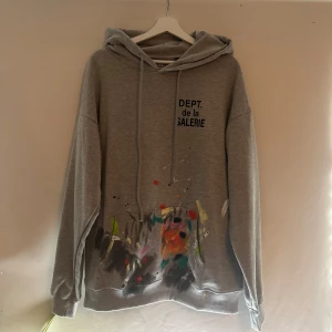 Grå hoodie från Dept de la Galerie - Säljer en grå hoodie från Dept de la Galerie med coola färgstänk på framsidan. Den har en avslappnad passform och är perfekt för både vardag och chill. Huvan är justerbar med snören och tröjan har långa ärmar. Perfekt för höst och vår!