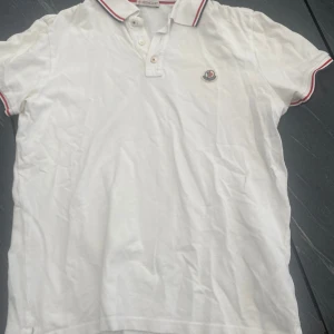 Vit piké Moncler - Vit piké t-shirt från Moncler med klassisk krage och knappar framtill. Röd och blå rand på krage och ärmslut samt broderad Moncler-logga på bröstet. Perfekt för en stilren och sportig look.