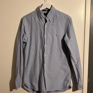 Ljusblå skjorta från Polo Ralph Lauren - Klassisk ljusblå skjorta från Polo Ralph Lauren med broderad logga på bröstet. Skjortan har lång ärm, knappar hela vägen och button-down krage. Perfekt för en clean och stilren look. Använd fåtal gånger.